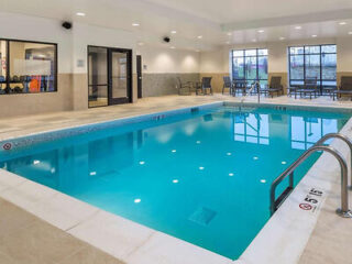 Indoor swimming pool with clear blue water, surrounded by beige tiles. Lounge chairs and a small table are visible near large windows in the background.