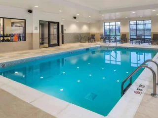 Indoor swimming pool with clear blue water, surrounded by beige tiles. Lounge chairs and a small table are visible near large windows in the background.