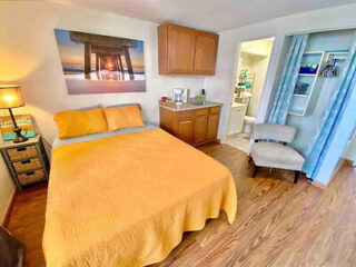 Bedroom with a yellow bedspread, wooden floor, bedside table with a lamp, chair, wall art, and cabinets. Adjacent bathroom with open door, visible sink, and shower curtain.