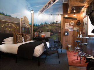 Rustic-style hotel room with western-themed decor, featuring a mural of a saloon and landscape, a bed with black and white bedding, a small table with chairs, and vintage signs on the walls.