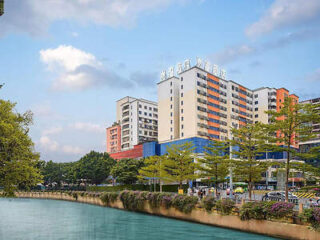 Riverside view of a modern multi-story building with colorful facades, surrounded by trees under a partly cloudy sky.