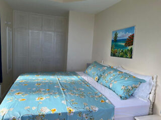 A bedroom with a bed covered in blue floral sheets, a painting of a tropical scene on the wall, and white closet doors in the background.