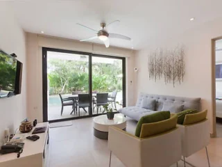 Living room with a sofa, two chairs, a TV on the wall, and a ceiling fan. Glass doors open to a patio with a table and chairs, overlooking a garden.