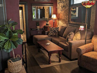 A cozy living room with a gray sofa, wooden coffee table, patterned armchair, and multiple lamps. Potted plants are placed in the corners, and artwork hangs on the walls.