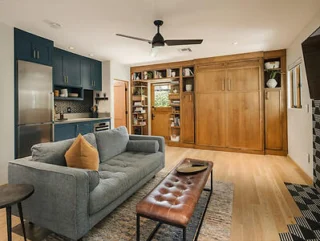 Open-plan living area with a gray sofa, brown leather bench, and a kitchen featuring dark cabinets and stainless steel appliances. Wall-mounted shelves and a ceiling fan are visible.