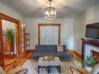 Living room with a gray sofa, a coffee table, and a wall-mounted TV above a fireplace. There's a chandelier, wood flooring, a plant, and a shelf with glass doors in the background.