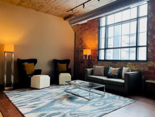 Loft-style living room with exposed brick, large window, two black armchairs, a grey sofa, modern lamps, and a patterned rug under a glass coffee table.