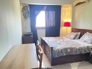 Cozy bedroom with a double bed, floral bedding, a wooden desk with a chair, a red floor lamp, a window with black curtains, and an air conditioning unit on the wall.