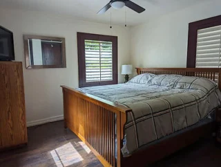 Bedroom with a wooden bed, striped bedding, nightstand with a lamp, wooden dresser, wall mirror, and window with shutters. Ceiling fan above.