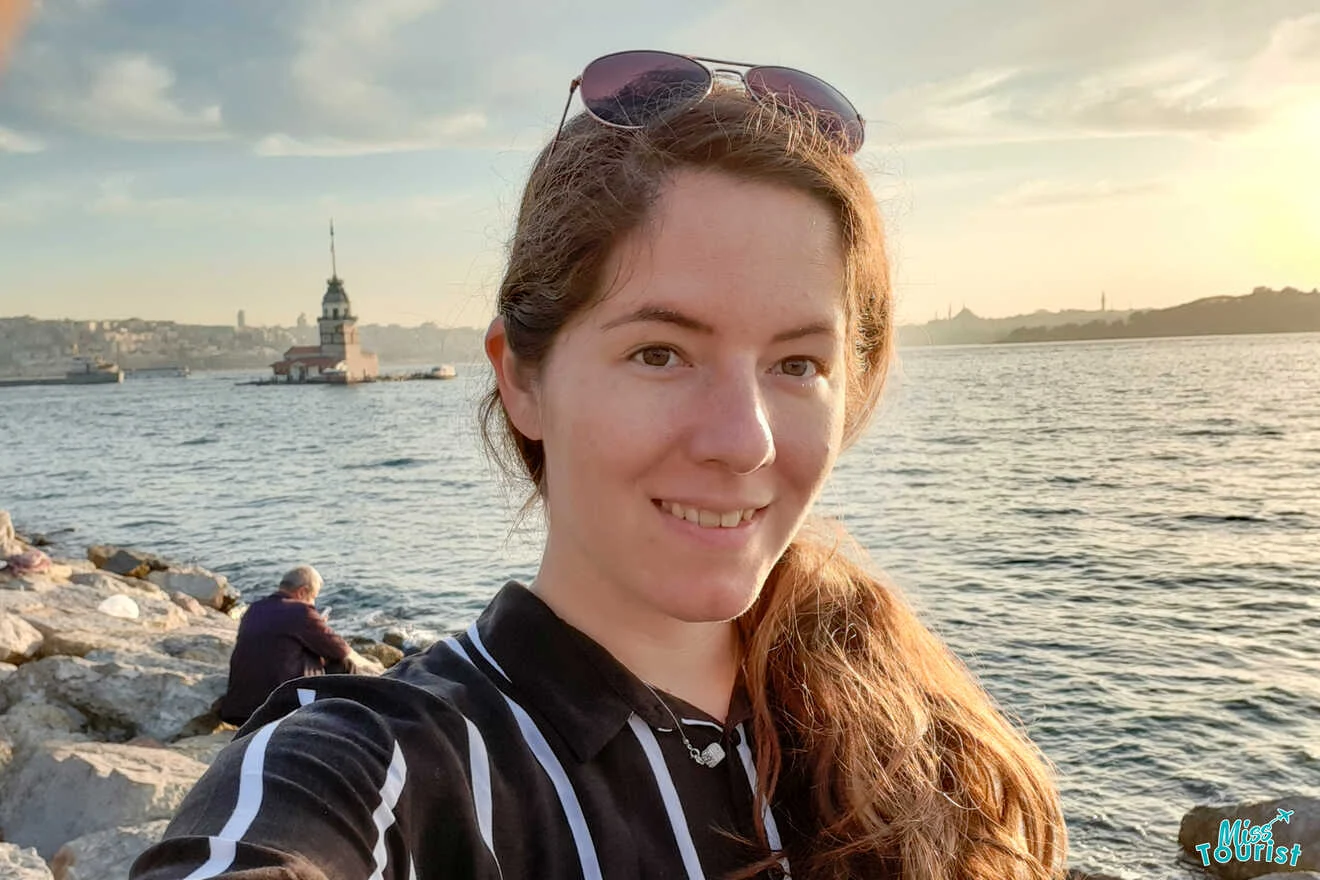 author of the post takes a selfie by a waterfront with a small tower on an island in the background.