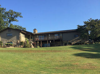 A single-story house with a stone chimney and wooden deck is situated on a grassy hill under a clear blue sky.