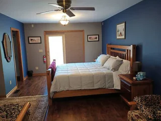 A bedroom with a wooden bed, white bedding, two nightstands, and paintings on blue walls. There's a ceiling fan, a large window with blinds, and a carpet on the wooden floor.