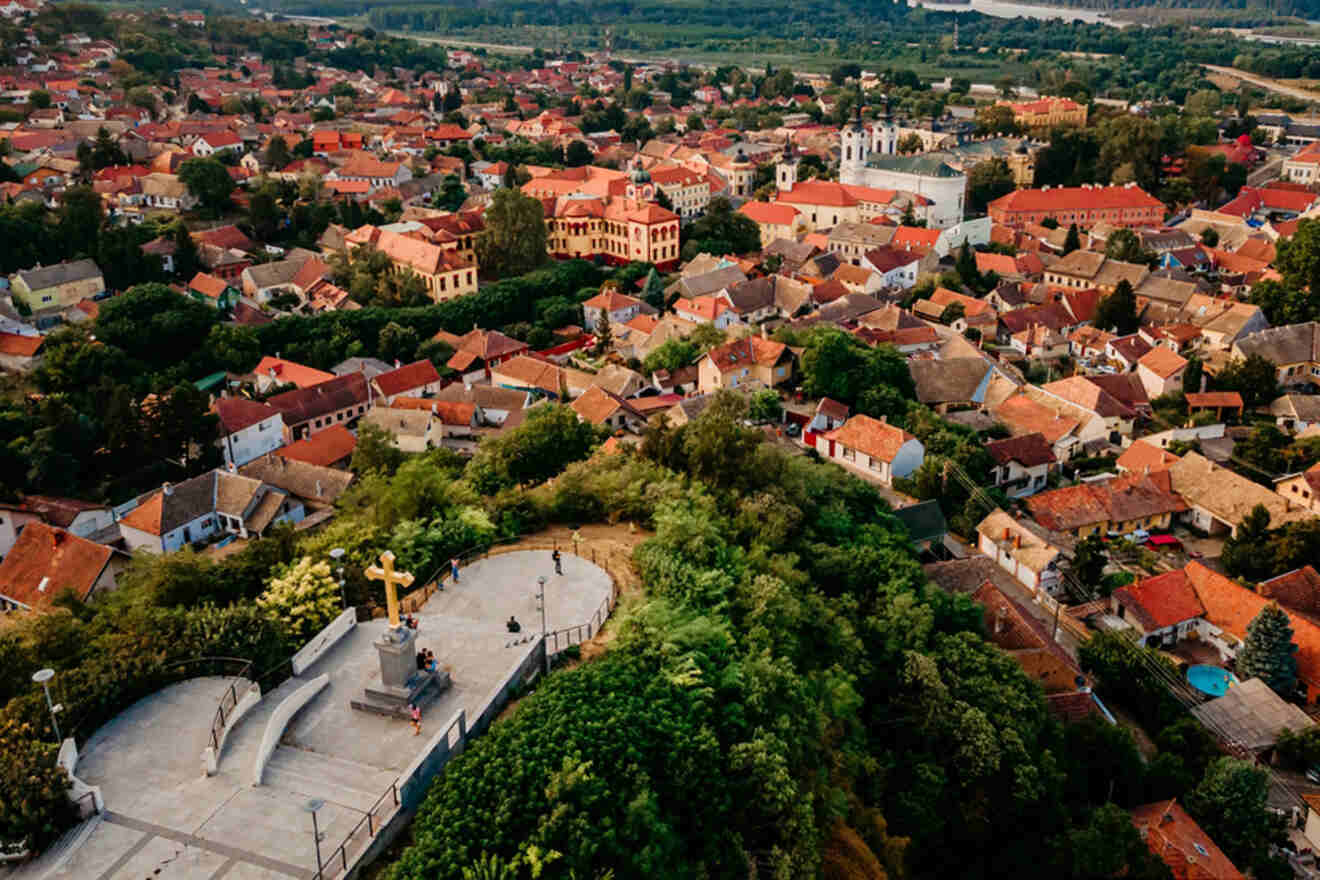 Aerial view of a town with red-roofed buildings, surrounded by greenery. A large cross and statue stand prominently in the foreground.