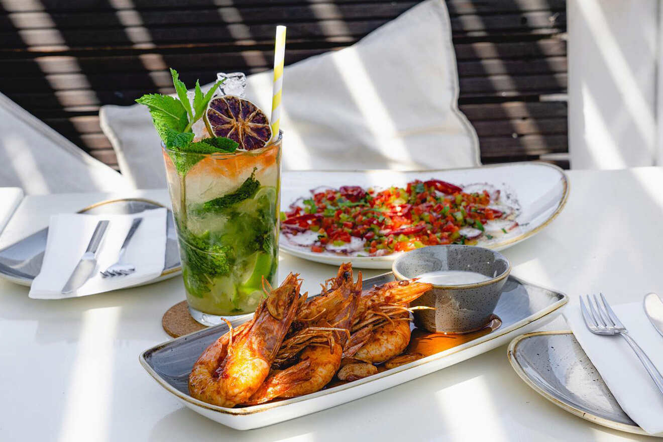 A table with a mojito, a plate of grilled shrimp with dipping sauce, and a plate of salad, set outdoors with striped shadows.