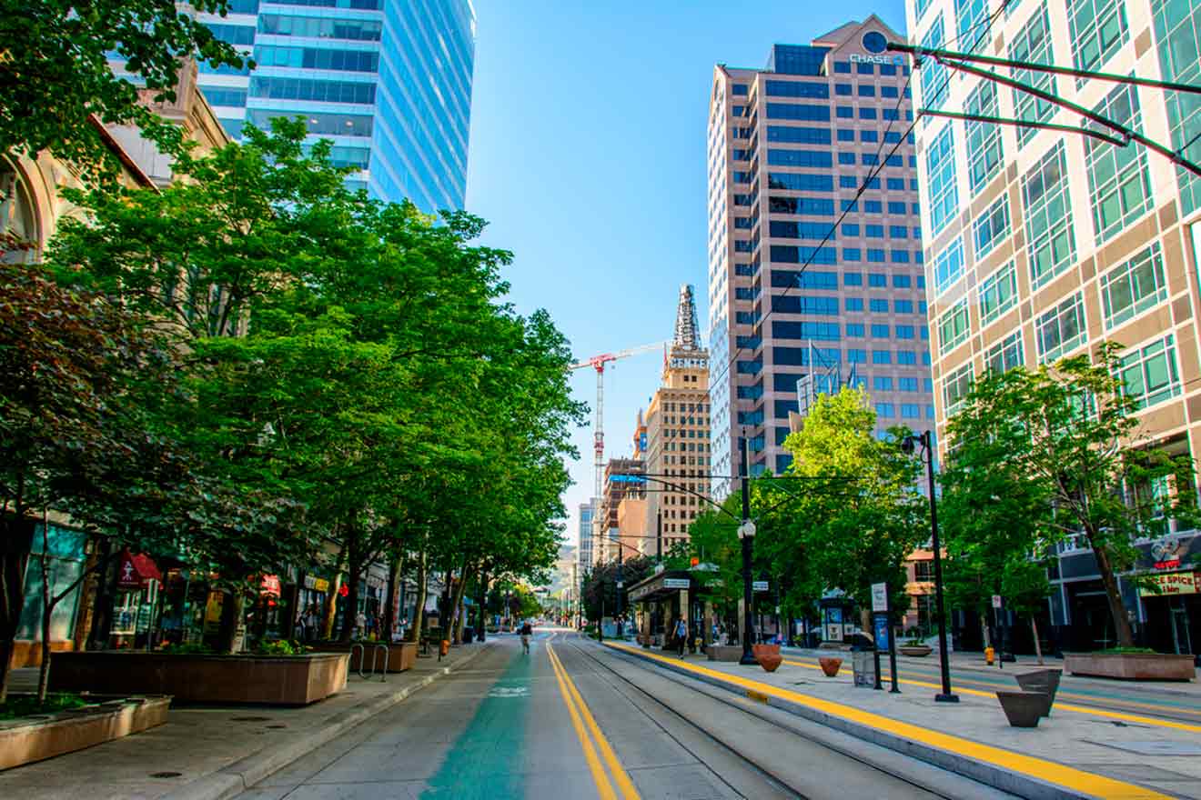 A city street lined with tall buildings and trees under a clear blue sky. A light rail track runs down the center.