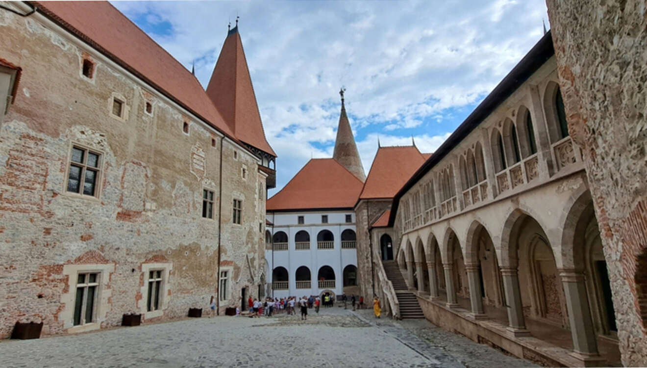 Medieval castle courtyard with tall spired towers, brick and stone walls. Tourists explore the cobblestone area under a partly cloudy sky.