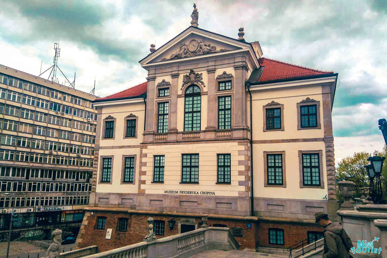 Historic building with classical architecture, featuring a red roof and ornate facade. Adjacent to a modern office building under a cloudy sky.