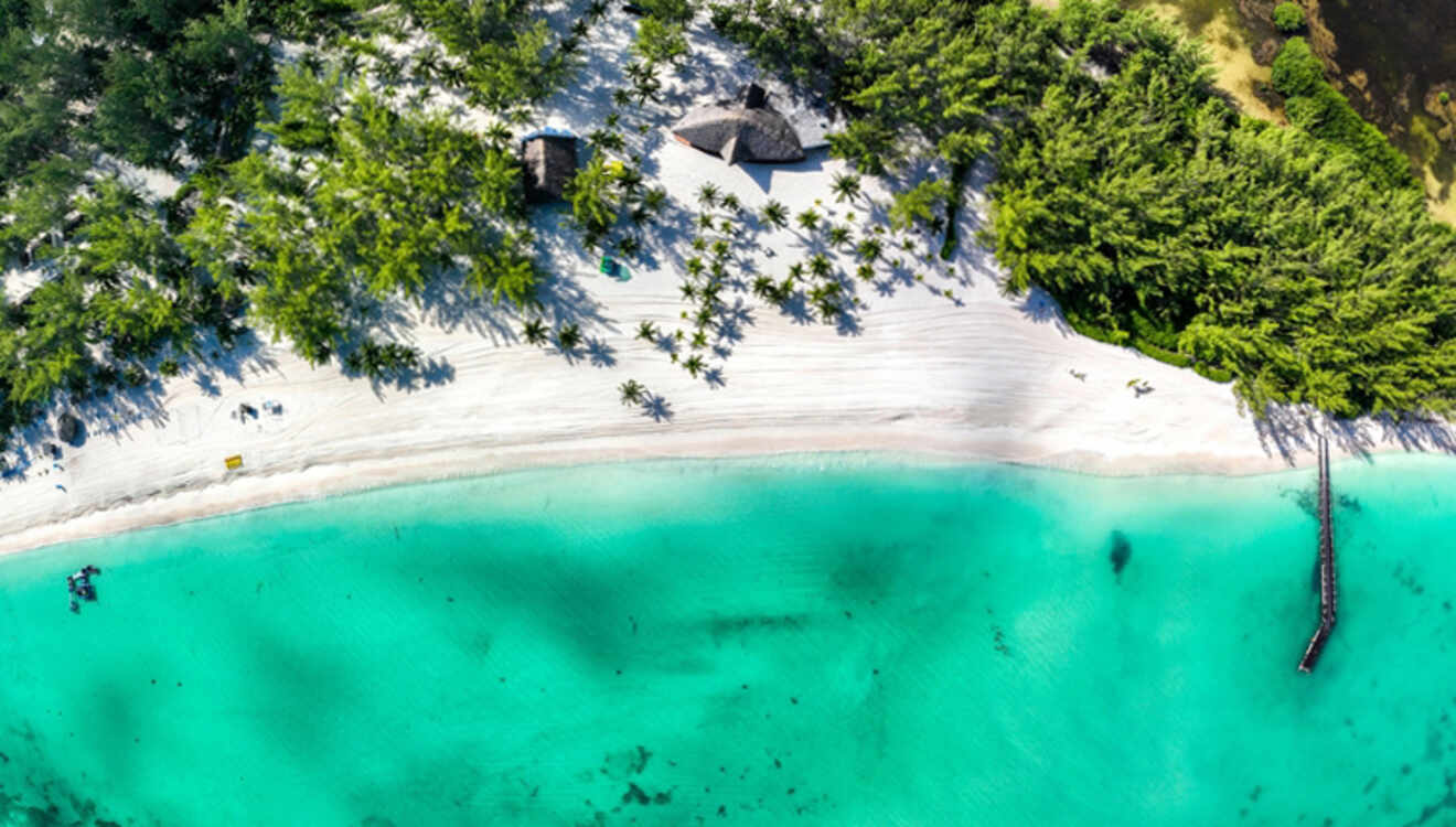 Aerial view of a tropical beach with turquoise water, white sand, palm trees, and a small pier extending into the sea.