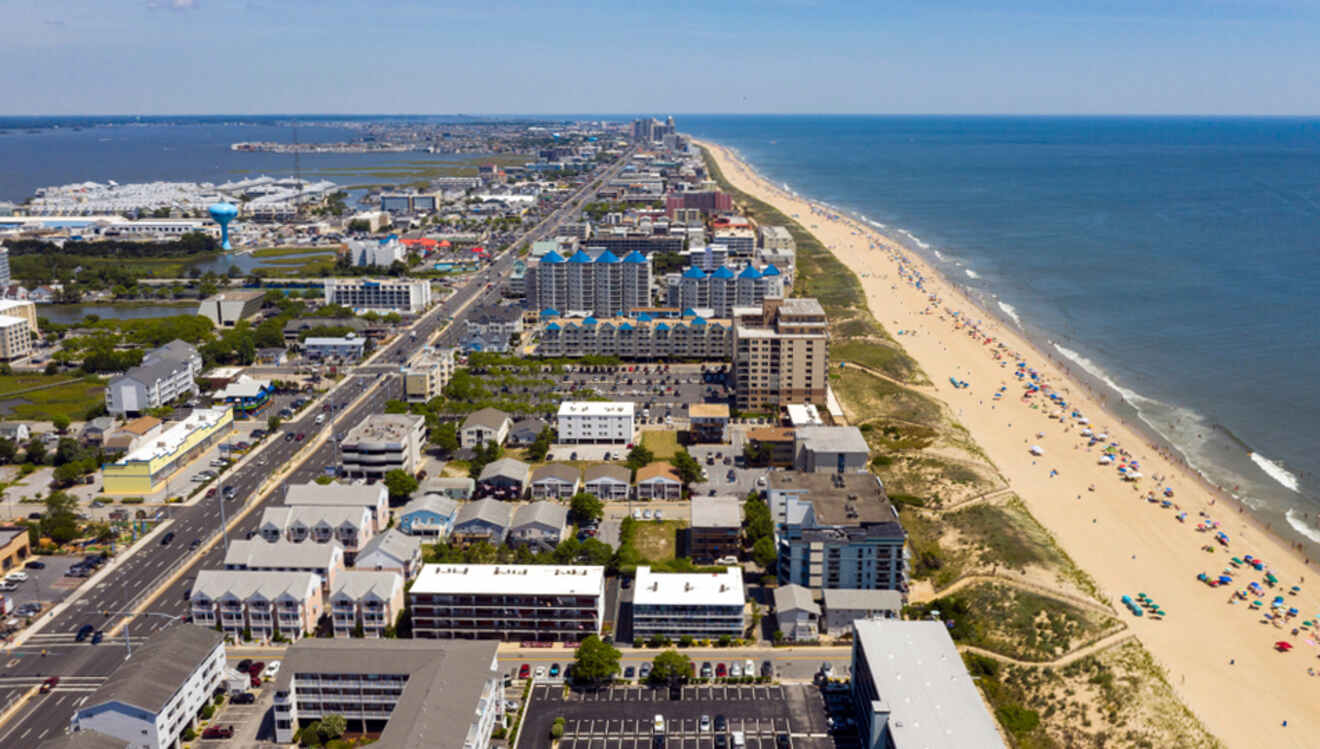 Aerial view of a coastal city with a sandy beach on the right, lined with colorful umbrellas, and urban buildings on the left.