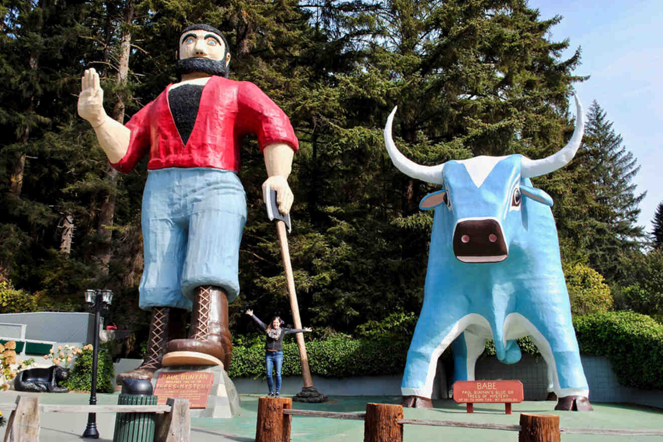 Giant statues of Paul Bunyan and Babe the Blue Ox stand in a forested area with a person posing between them.