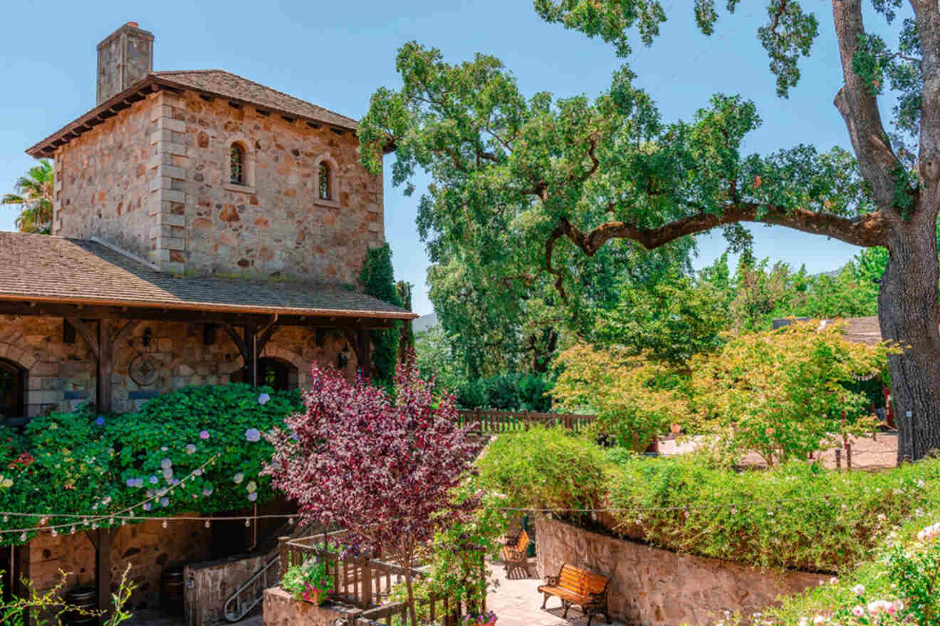 A stone building with a wooden roof is surrounded by lush greenery, trees, and colorful shrubs in a garden setting under a clear blue sky.