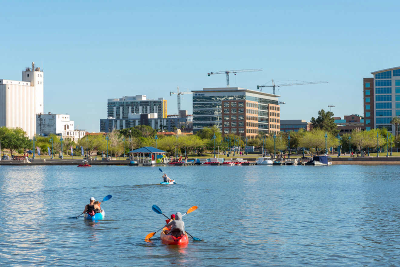 People kayaking on a calm river with a cityscape and construction cranes in the background.