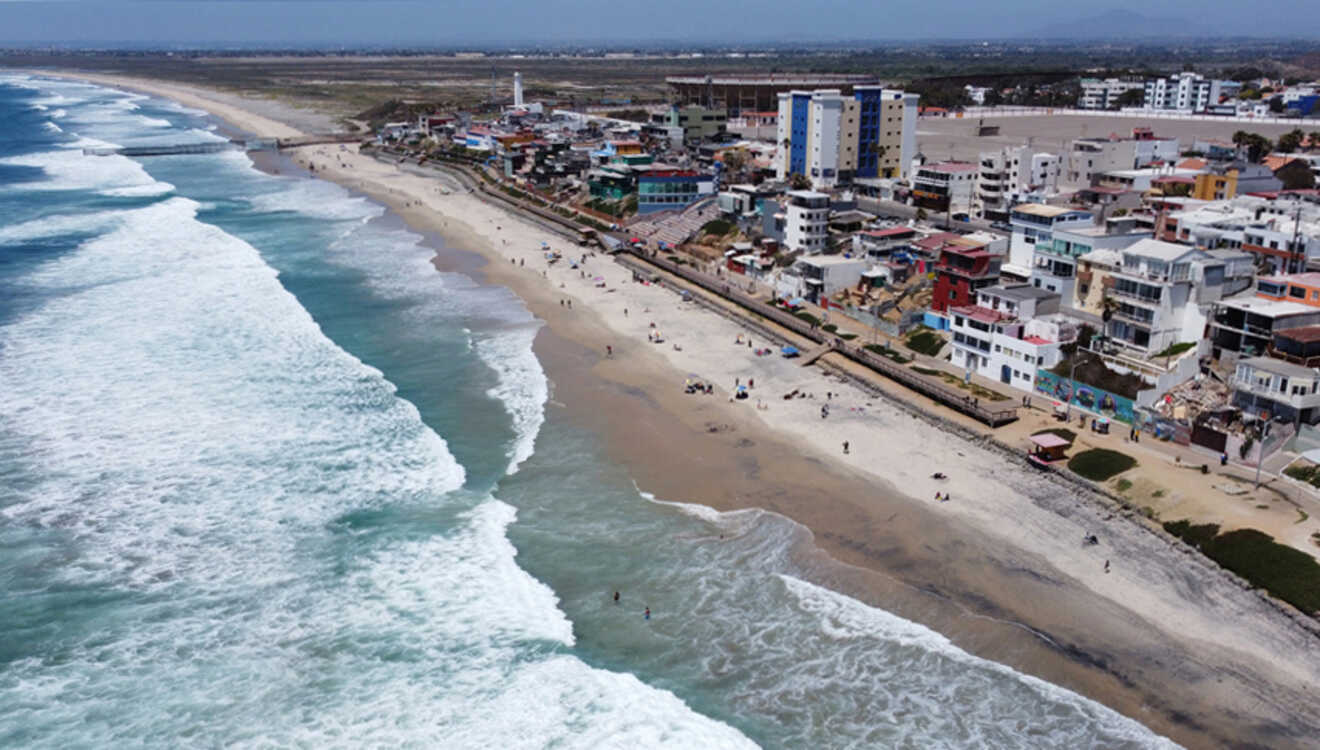 Aerial view of a sandy beach with waves crashing, bordered by a coastal town with colorful buildings and roads. People are walking along the shore and engaging in beach activities.