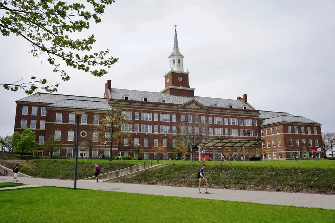 A large, historic brick university building with a central tower, bordered by green lawns and a pathway. People are walking in front of the building.