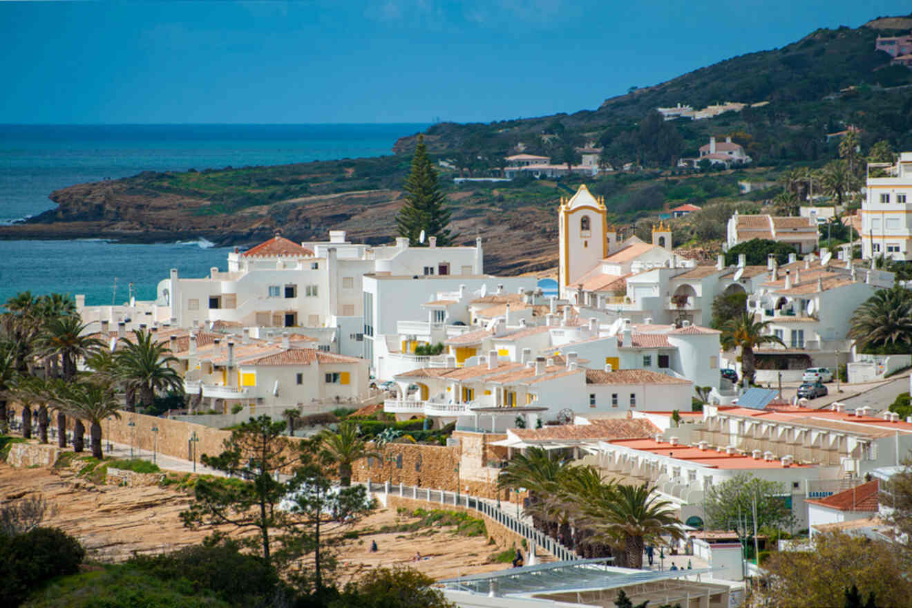 Nestled in a coastal town, white buildings and palm trees create a serene landscape, with the church standing proudly against the backdrop of hills and the shimmering sea, reminiscent of picturesque Lagos, Portugal.