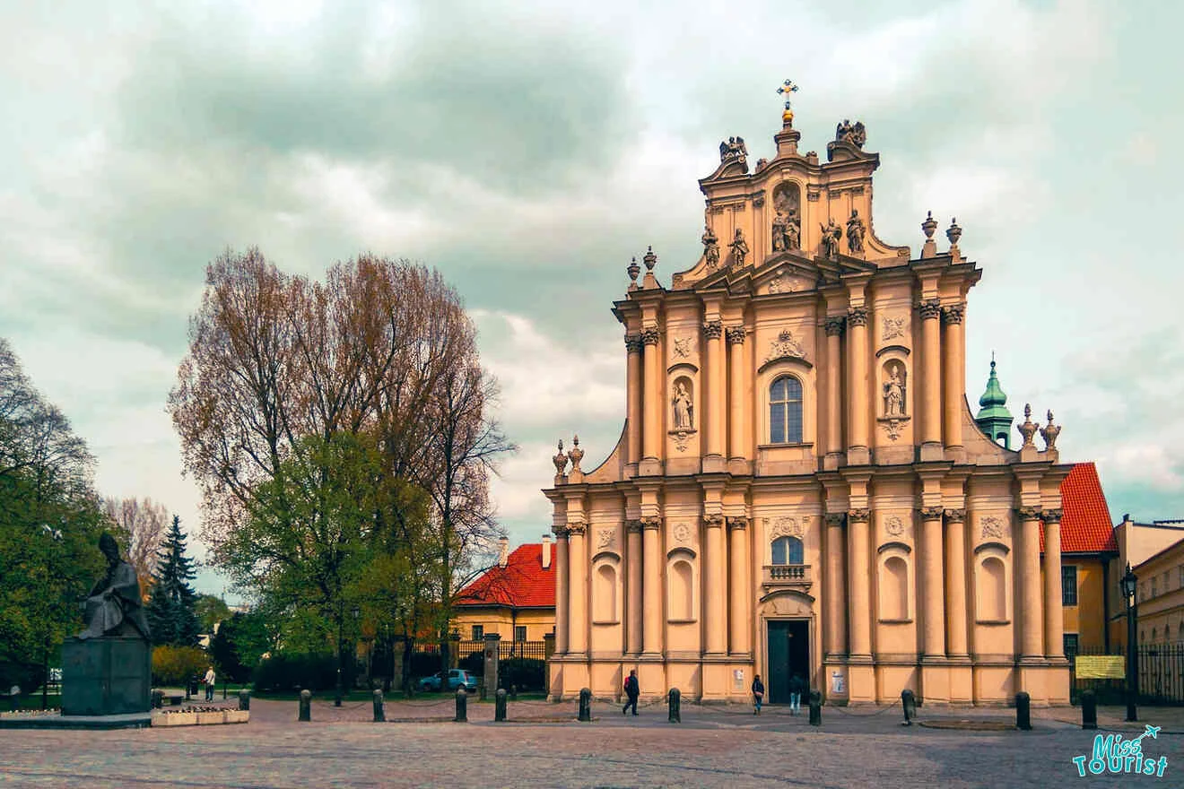 Baroque-style church with ornate facade and statues, situated on a square with trees and a statue, under a cloudy sky.