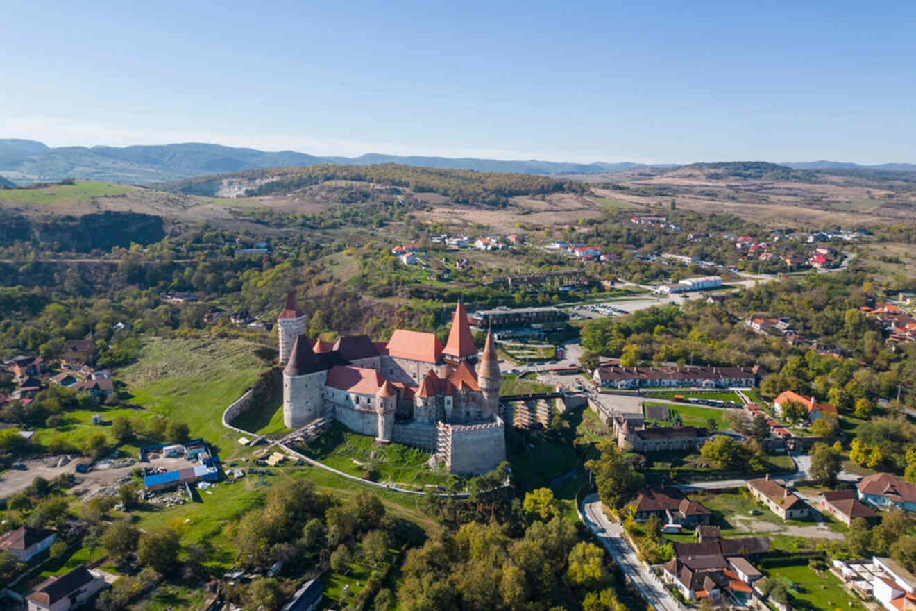 Aerial view of a historic castle with red roofs surrounded by lush greenery, small houses, and rolling hills under a clear blue sky.