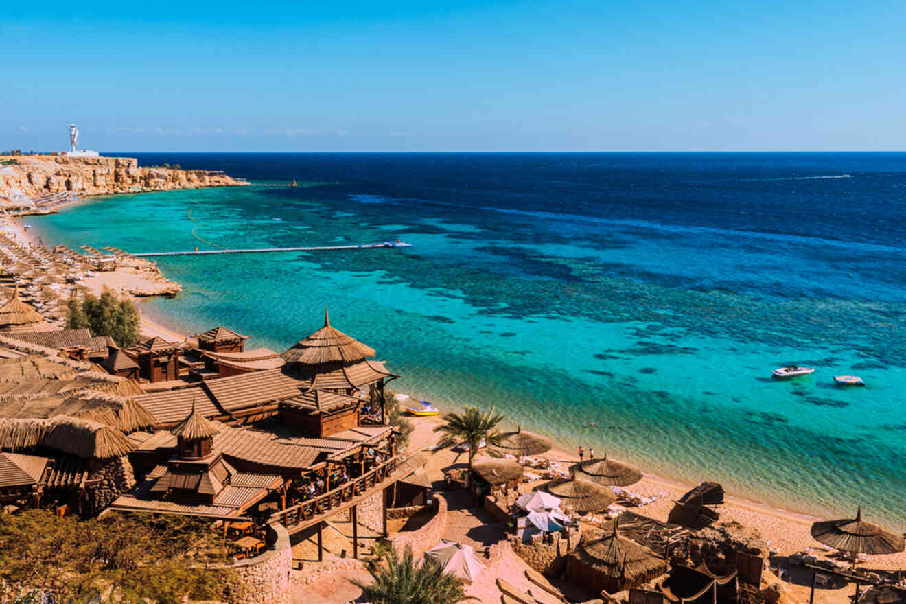 A scenic coastal view with turquoise water, sandy beach, and wooden huts with thatched roofs under a clear blue sky.