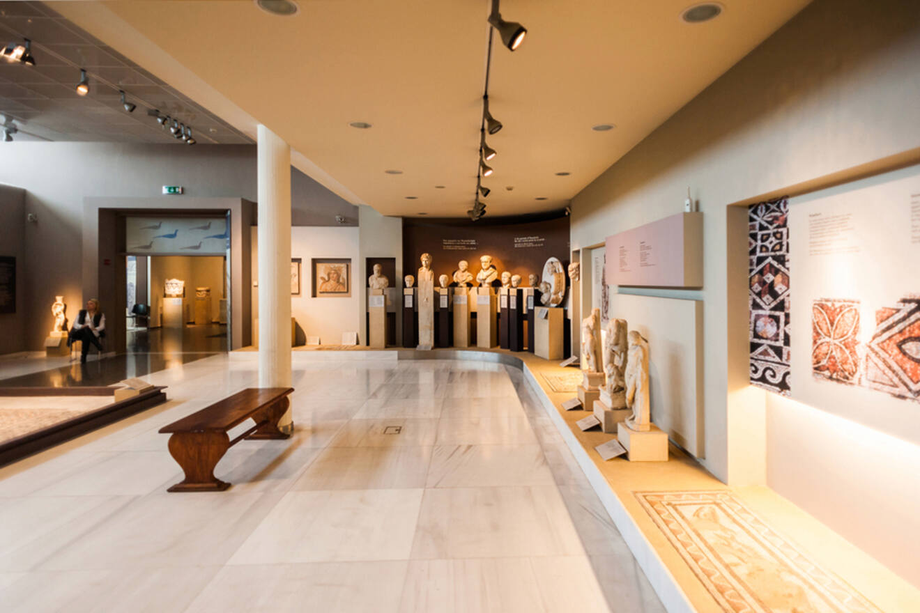 Interior view of a museum gallery with sculptures on display, informational panels, and a wooden bench in the foreground. The floor is tiled, and track lighting illuminates the exhibits.