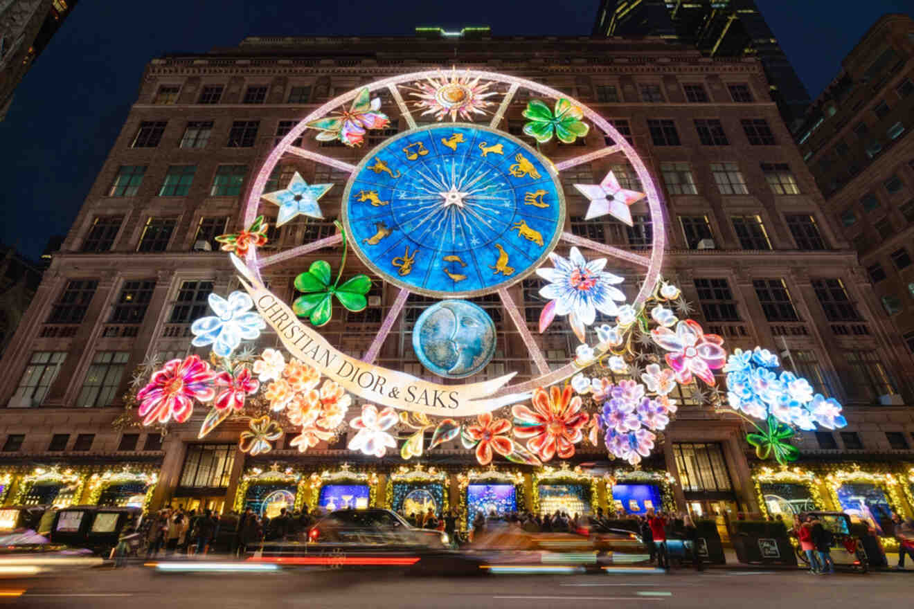 A building facade decorated with colorful, illuminated floral and zodiac displays. Text reads "Christian Dior & Saks." Busy street with moving cars in the foreground.