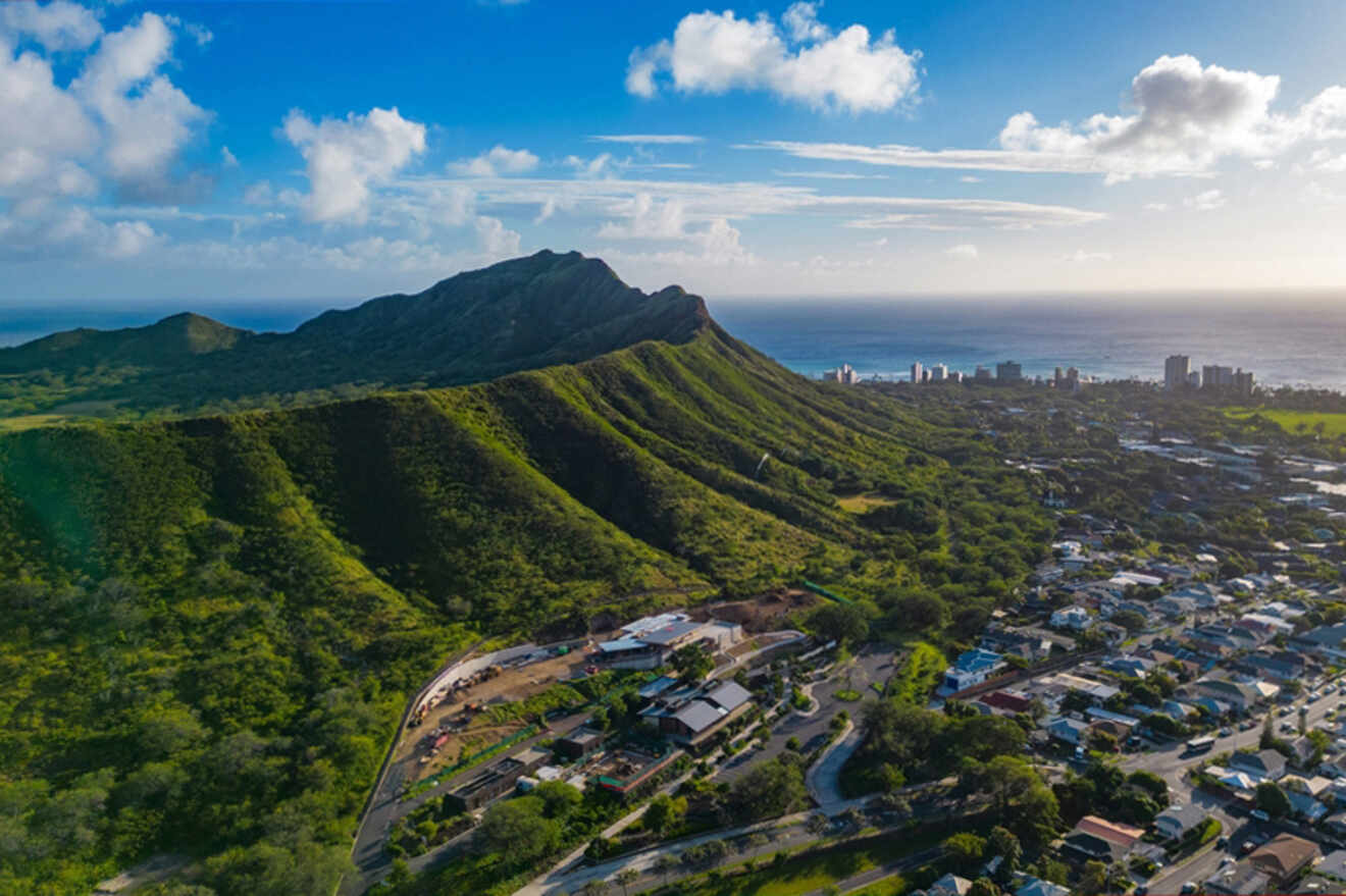 Aerial view of a lush green mountain, possibly Diamond Head, with a cityscape and ocean in the background under a partly cloudy sky.