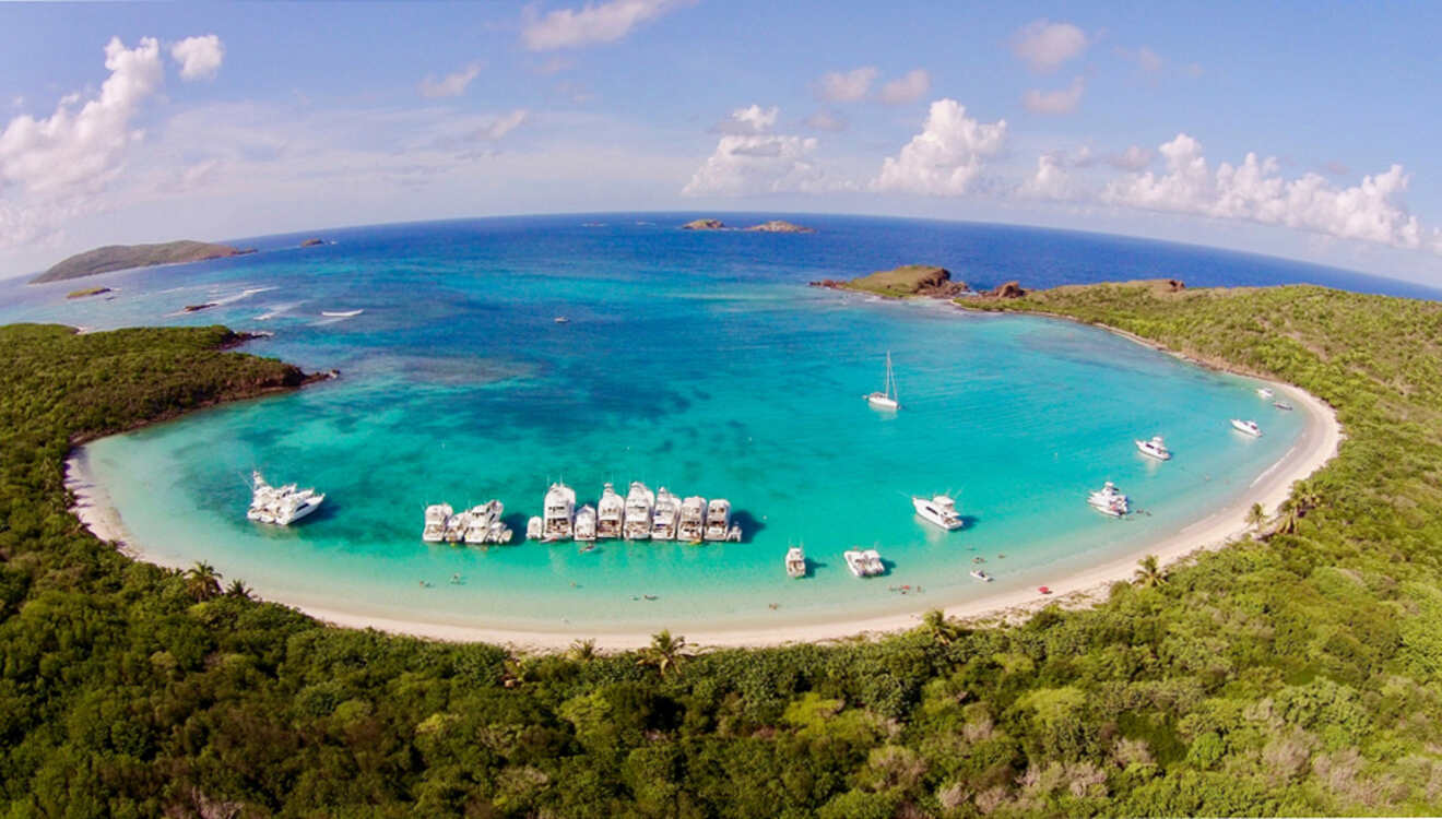 Aerial view of a circular turquoise bay with several boats anchored near a sandy beach, surrounded by lush green landscape and open sea under a blue sky.