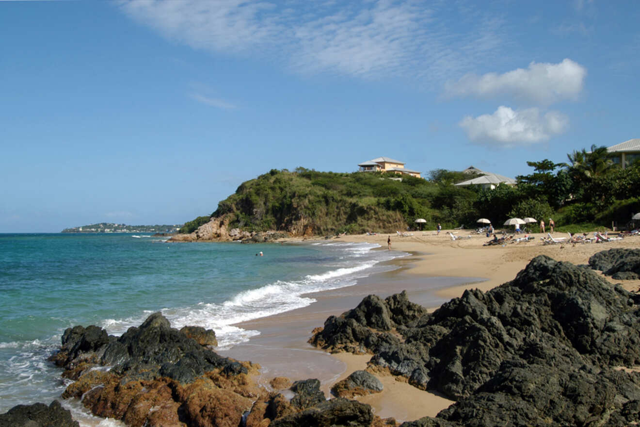 A rocky shoreline with a sandy beach, clear blue water, and a few houses in the background under a blue sky with scattered clouds.