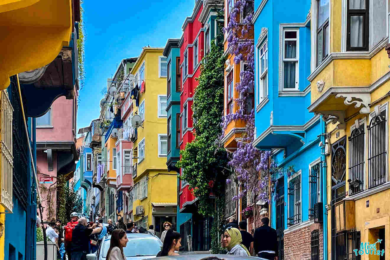 A vibrant street with colorful buildings, people walking, and cars parked. Purple flowers climb one building.