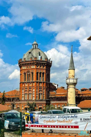 A historic brick building with a domed roof stands beside a slender minaret. Banners and vehicles are in the foreground under a blue sky with clouds.