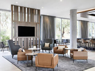 Modern lounge area with brown chairs, gray armchairs, a large TV on a wooden panel, and floor-to-ceiling windows.