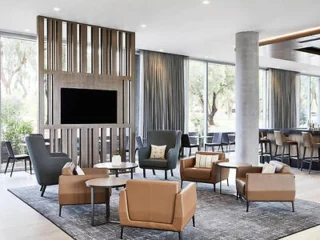 Modern lounge area with brown chairs, gray armchairs, a large TV on a wooden panel, and floor-to-ceiling windows.