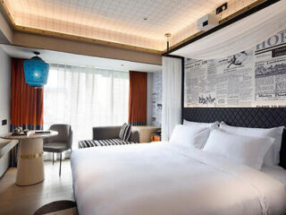 Modern hotel room with a large bed, a colorful lamp, and wall art featuring newspaper prints. There's a small table with chairs near a window with orange curtains.