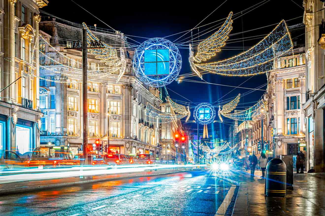 A busy city street at night with ornate holiday lights, including glowing spheres and wing shapes, strung between historic buildings. Traffic creates light trails on the wet pavement.