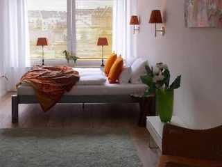 A cozy bedroom with a neatly made bed, orange pillows, and a warm throw. Two wall lamps flank the window, and a vase of flowers sits on a white table. Green rug on the wooden floor.