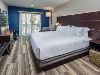 Hotel room with a large bed, striped carpet, desk, chair, and a sliding glass door leading to a balcony.