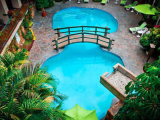 Aerial view of a swimming pool with a bridge across two sections, surrounded by lush greenery and lounge chairs.