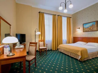 A hotel room with a double bed, two chairs, a desk with a lamp, a TV, and large windows with yellow curtains. The floor is carpeted, and a painting hangs on the wall.