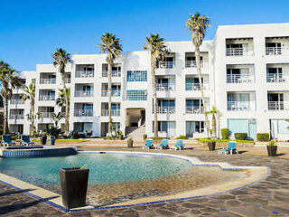 Modern white four-story building with balconies, surrounded by palm trees, faces a circular pool with lounge chairs on a stone patio.