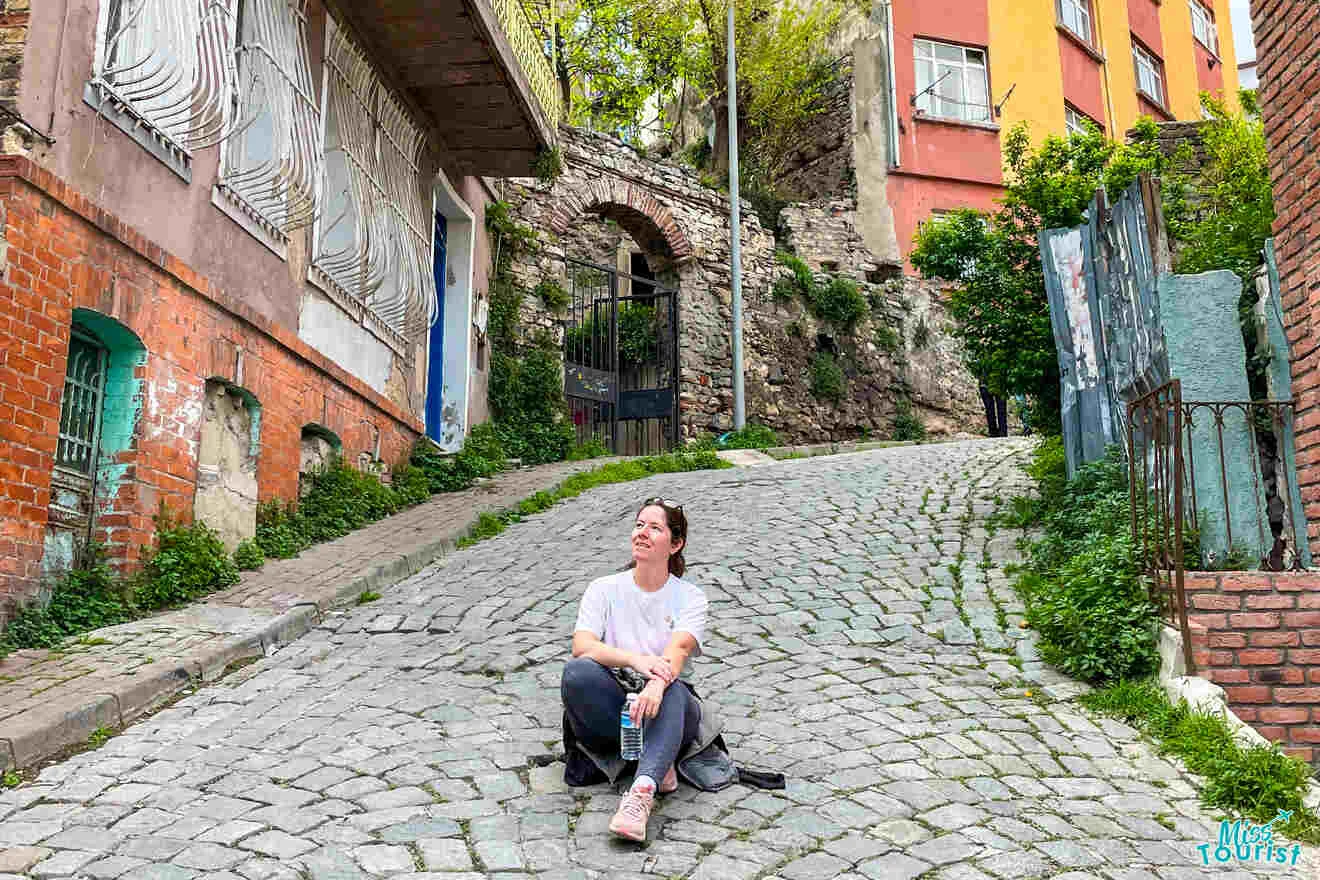 author of the post sitting on a cobblestone street, holding a water bottle, with colorful buildings and greenery in the background.