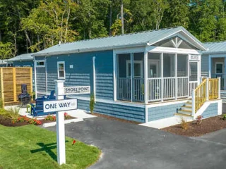 Small blue cottage with a screened porch, surrounded by greenery. A "One Way" street sign is visible in the foreground.